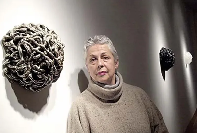 A light-skinned adult woman with short, grey hair. She gazes directly into the camera with a frank expression, her eyebrows slightly raised. On the wall next to her hang three round sculptures of grey, black and white. They are masses of coils and resemble rounded brains.