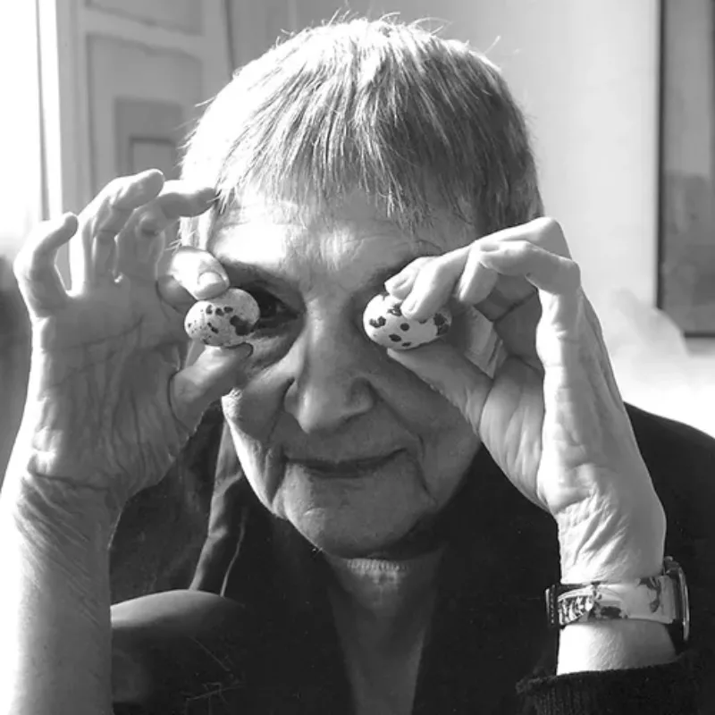 Close-up black and white photo of an older woman with light skin and cropped hair. She holds up two small, speckled eggs to her eyes, the right one just visible. She sits in a bright, sun-lit room, a window on her right and wears a dark blazer and wrist watch.