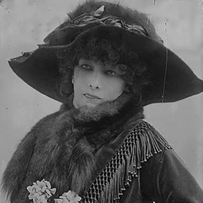 Black-and-white photo of a light-skinned adult woman with dark curls. Starring off-camera, she wears a large dark hat, a dark dress with long sleeves, and fur stole. Her mouth opens slightly, showing her top row of teeth, and she holds a bouquet, visible at the bottom of the frame.