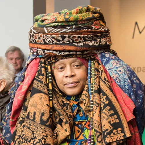 A medium skin-toned adult woman smiles with only her face visible from underneath an elaborate headwrap of nearly a dozen different colorfully printed fabrics. The wrap is piled like a turbin atop her head with fabric cascading down to surround her face.