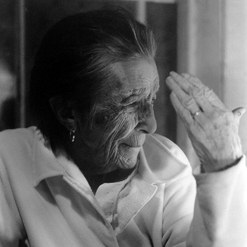 Black-and-white photograph of an older woman with her short, dark hair brushed back. Sitting with her left hand raised, her deeply lined face looks off-camera, her eyes squinting slightly and her lips pursed. She wears a gold earring, gold wedding ring and a white shirt.
