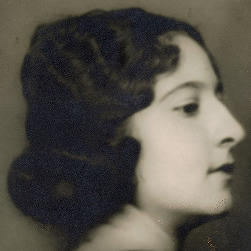 Soft-focus black-and-white studio portrait of the head of a light-skinned young adult woman in profile. Her curly dark hair is pulled back in a low bun.