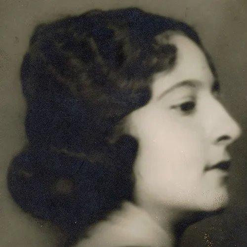 Soft-focus black-and-white studio portrait of the head of a light-skinned young adult woman in profile. Her curly dark hair is pulled back in a low bun.