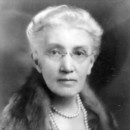 A black-and-white soft-focus studio portrait photograph of an older light-skinned woman with white hair pulled back. She wears rimless glasses, three strands of pearls around her neck, and a fur wrap over her shoulders.