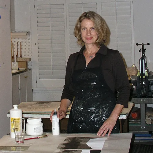 A light-skinned adult woman with shoulder length blond hair. She wears a paint covered apron and stands behind a tablle covered in art supplies, incluidng brushes and photographs. She smiles slightly, her hands posed as if they were about to begin creating.