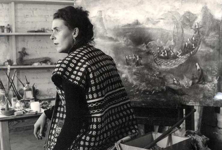 Black-and-white photograph of a light-skinned adult woman with dark curly hair. She sits with her back to the viewer, looking over her left shoulder, her face in profile, surrounded by art supplies. A painting of nuns in a boat on rough seas sits on an easel in front of her.