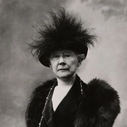 Black-and-white photograph of a seated older woman with light skin. She wears a dark dress with a fur stole and long necklace. Her dark hat is adorned with a group of large, wispy feathers and covers her hair. Her expression is open and calm.