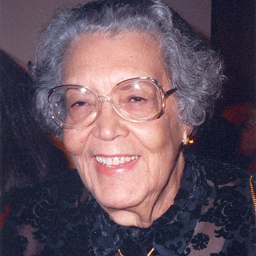 A medium skin-toned older woman with short gray hair smiles at the viewer. She wears clear-framed glasses and a black, floral patterned shirt.