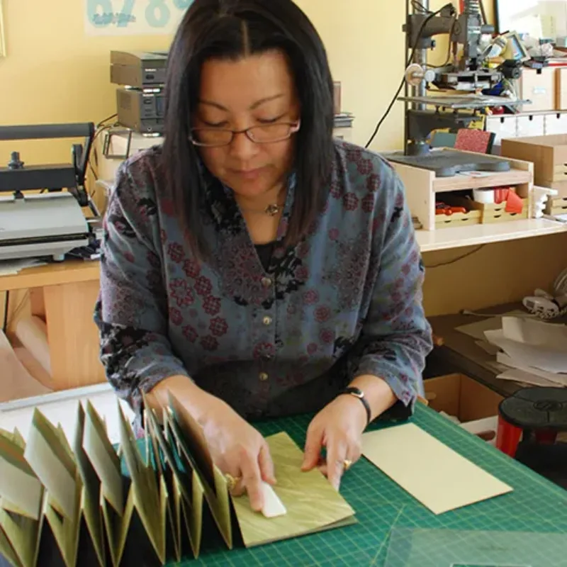 A light-medium-skinned adult woman with medium-length black hair wears a blue floral blouse and glasses. She is in an art studio, behind a green cutting mat, looking down and holding open a flag-style book. Equipment is on the counter behind her.