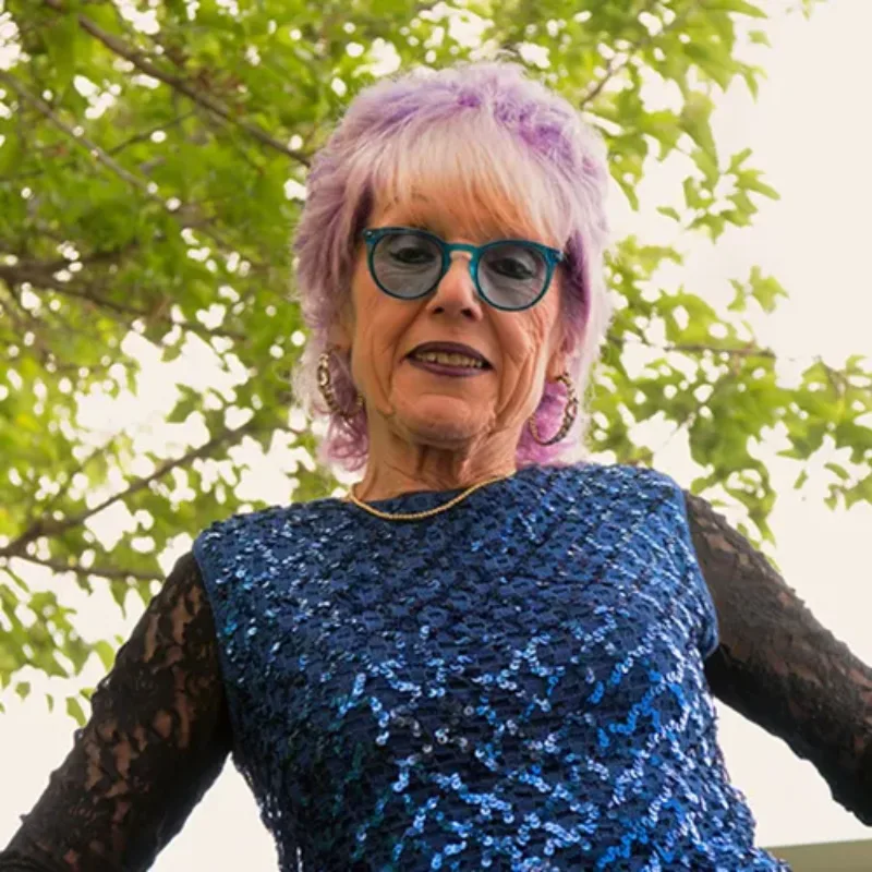 A light-skinned older woman with short purple-and-white hair smiles slightly, arms out to the side. She wears a blue sequin top with long black lace sleeves, a gold necklace, hoop earrings, and blue glasses. The sky and tree branches are behind her.