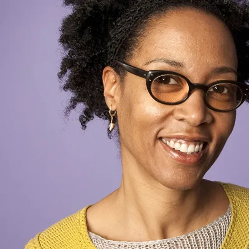 A woman with medium-dark skin tone and short black hair smiles into the camera. She wears black glasses, a white and yellow knit top and gold earrings.