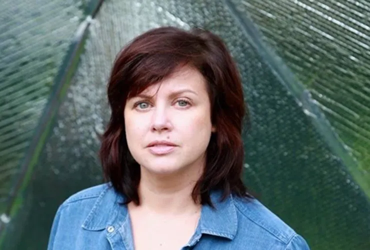 A light-skinned adult woman seen from the shoulders up. Her dark hair reaches to her shoulders, clad in denim. She has green eyes and a mole above the left side of her mouth. Her green eyes look straight into the camera and match the green behind her.