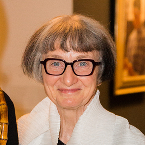 A light-skinned older woman with dark hair streaked with gray, cut in a short bob. She wears a white, high-colored top. She smiles as her blue-gray eyes gaze directly at the camera from behind eyeglasses with a thick, dark frame.