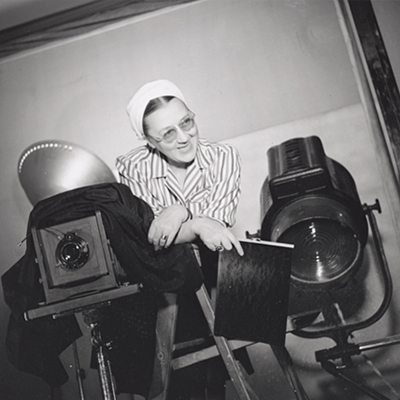 Black-and-white photo of a light-skinned adult woman leaning out from behind a large studio camera. Wearing glasses, a white turban and a stripped top, she smiles with an expression of humor. A large spotlight sits behind her and she holds a piece of large format film.