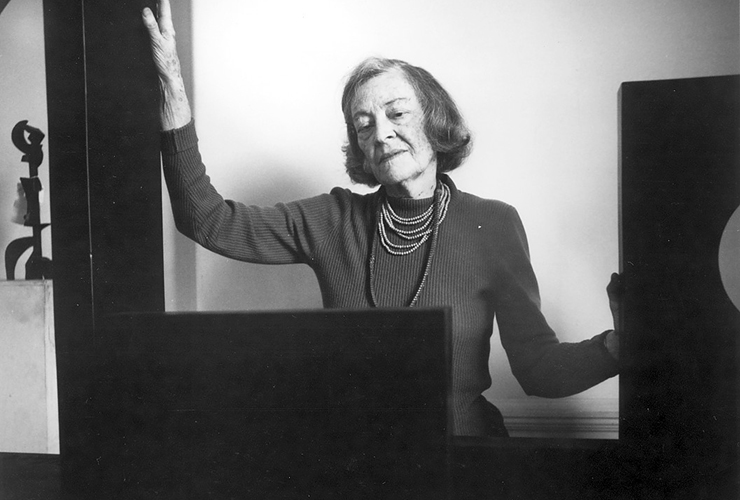 A black-and-white photograph of a light-skinned older woman with short hair stands looking at a black rectangular sculpture. She wears a dark long sleeved shirt and several beaded necklaces. Both her arms are outstretched as she touches other black sculptures beside her.