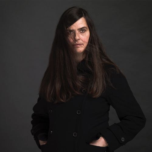 A light-skinned adult woman wearing a dark jacket against a dark background. Her hands are tucked into pockets. Her long dark brown hair falls across her forehead, partially obscuring her long eyebrows.