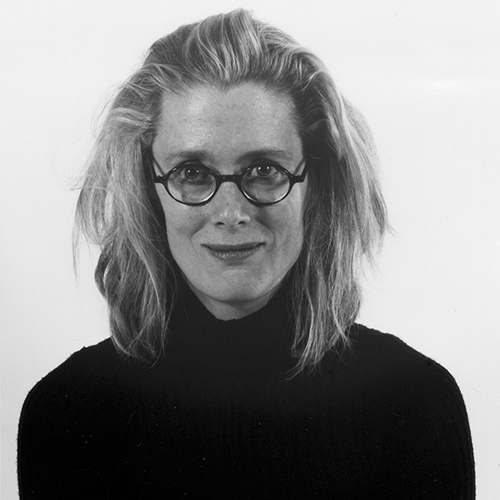 Black-and-white studio portrait of a smiling light-skinned adult woman with medium-length blond hair. She wears round glasses and a black turtleneck sweater.