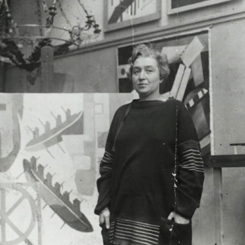 A black-and-white photograph of a light-skinned, adult woman standing in her studio and surrounded by her large, abstract, geometric paintings. Her hair is short, light, and curly, and her hands rest in the pockets of the loosely fitted, dark colored, long-sleeved dress she is wearing.