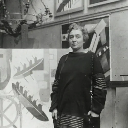 A black-and-white photograph of a light-skinned, adult woman standing in her studio and surrounded by her large, abstract, geometric paintings. Her hair is short, light, and curly, and her hands rest in the pockets of the loosely fitted, dark colored, long-sleeved dress she is wearing.