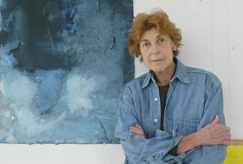 A light-skinned older woman with short, wavy, red hair and brown eyes stands with her arms crossed. She leans against a white wall with part of a blue abstract painting behind her. She wears round silver-color earrings and a blue denim long-sleeve button-up shirt.