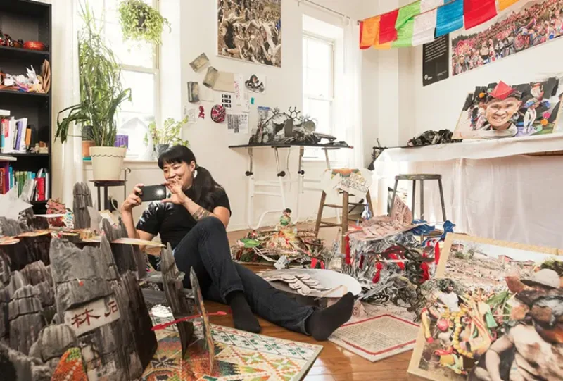 A light-skinned, adult, Asian woman with black hair dressed in black sits on the floor of her cluttered studio surrounded by elaborate pop-up books. She holds up her mobile phone to take a photo.