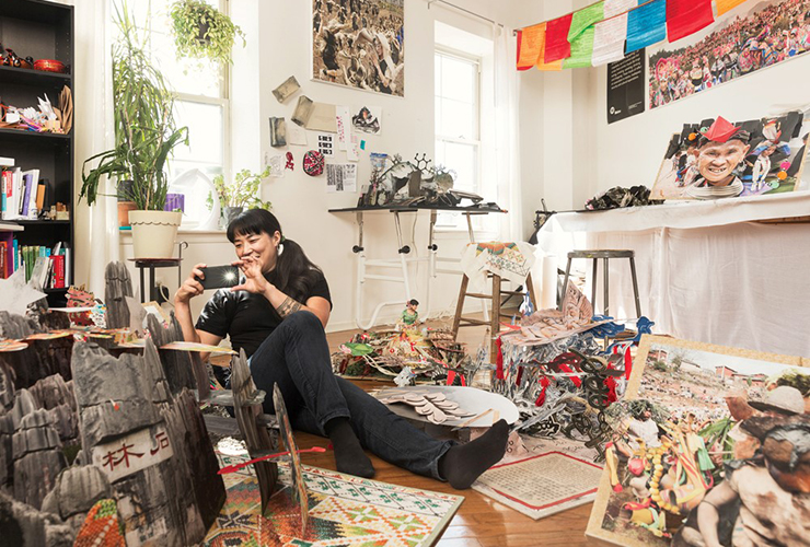 A light-skinned, adult, Asian woman with black hair dressed in black sits on the floor of her cluttered studio surrounded by elaborate pop-up books. She holds up her mobile phone to take a photo.