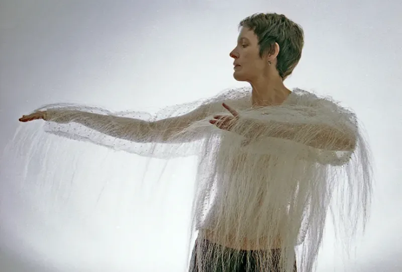 A light-skinned adult woman with short brown hair stands in front of a blank wall. Her head is turned to her right, her right arm is stretched out, and her left arm is bent in front of her chest. She wears a sheer white long-sleeve blouse with long white hanging threads.