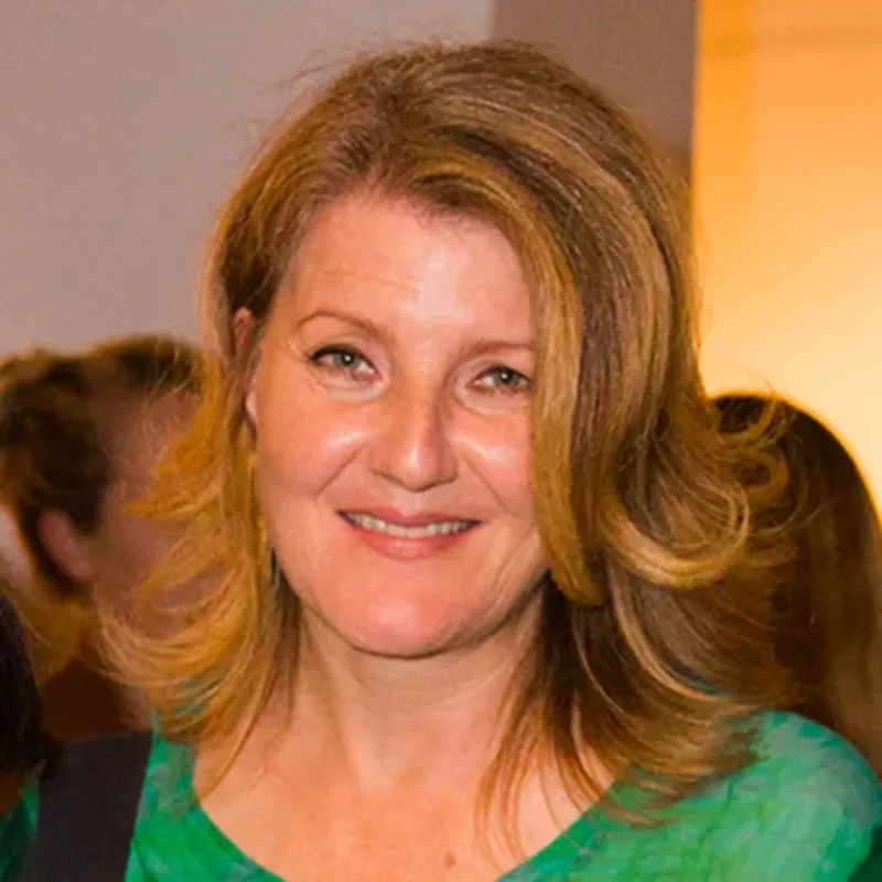 A light-skinned adult woman with slightly curled blonde hair and a green top smiles while standing in front of several people in shadow.