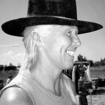 A black-and-white photograph of a light-skinned, smiling adult woman in profile wearing a black Western-style hat and tank top, her white hair pulled to the side. She stands in a field of sparse grass. Behind her are two people, a rack of clothing, furniture, and distant trees.