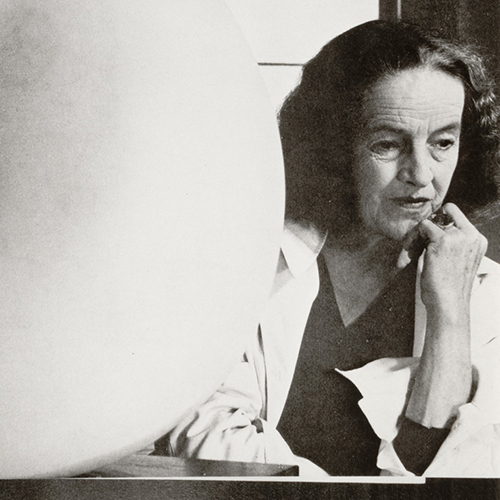 A black-and-white photograph of a light-skinned older woman with short, dark, wavy hair. She sits thoughtfully beside one of her large, white, round, abstract sculptures with her chin resting on one hand