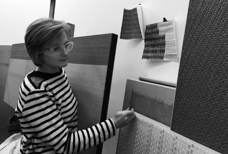 A black-and-white photograph of a light-skinned adult woman with short hair, stylish glasses, and a striped top, contemplating her many highly textured paintings that resembled magnified textile swatches. She thoughtfully holds a painting knife near one painting.