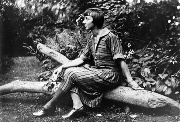 A black-and-white photograph of a light-skinned adult woman. Seated outdoors on a log in front of greenery, she is posed in profile with her body turned to the left of the composition. She wears a long striped dress and crosses her legs. She has short, dark hair cut in a bob.