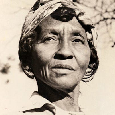 A black-and-white photograph of a dark-skinned older woman, shown from the neck up. She has short, dark hair, held away from her face by a white head wrap. She has a stoic expression, and is looking away from the camera against a light background.