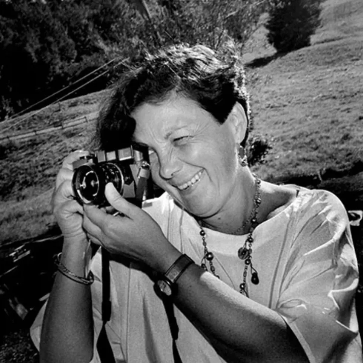 A black-and-white photograph of an adult woman with short, dark hair looking into the viewfinder of a small film camera. She is outdoors with grass behind her. She holds the camera with her right hand and focuses the lense with her left. She is smiling as she looks into it.