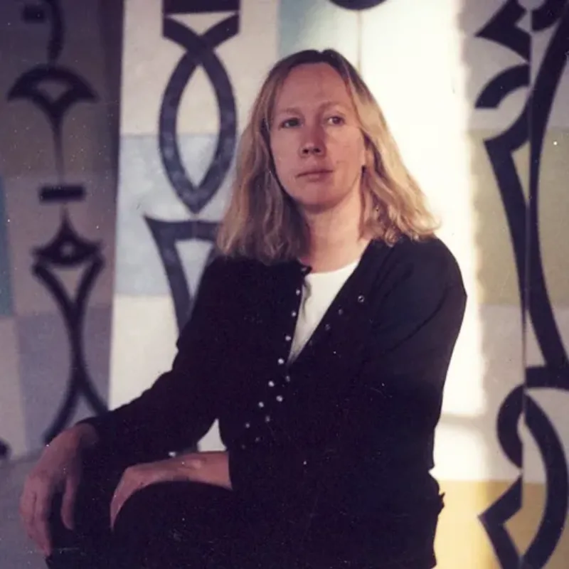 Color photo of a seated woman with her hands on her knees. She has shoulder-length blonde hair and light skin and looks past the camera. Behind her, abstract paintings on panels of yellow, white and blue covered in black symbols. A patch of sunlight lights the left side of her face.