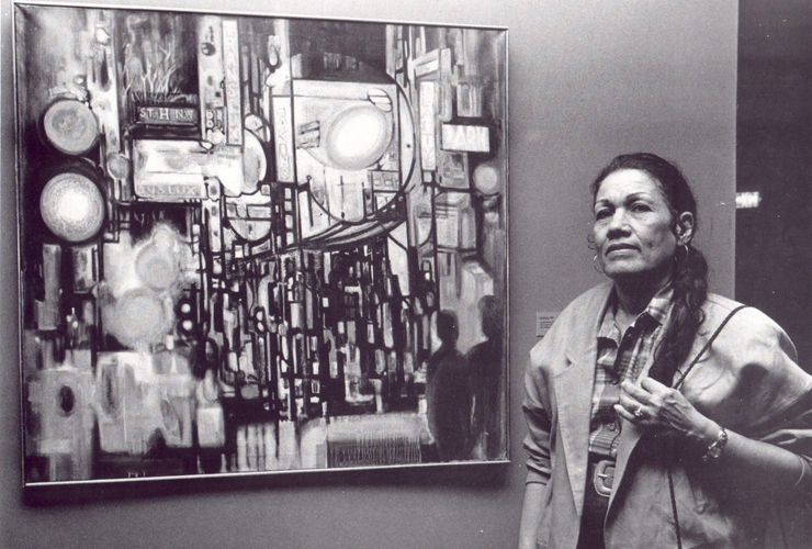 A black-and-white photograph of an adult woman with long dark hair in a braid. She wears a plaid shirt, jacket, large belt and hoop earrings. Her hand is touching her braid. She stands next to a painting which depicts an abstract street scene.