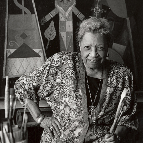 Black-and-white photograph of a smiling medium-skinned older woman with white curly hair. She wears a patterned blouse and necklace with figure pendant. She holds brushes in her left hand and rests her right hand on her hip. Behind her is a painting with a figure and shapes.