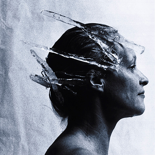 Close-up black-and-white studio portrait of a light-skinned adult woman in profile. Her hair is pulled back and she wears a crown of icicles. Small streams of liquid run down her face and neck.