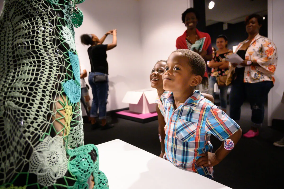 Two young children with medium skin tone stand in front of a dog sculpture covered with green crochet panels. One child stands with hands on hips, eyes turned to look directly at the viewer, and gives a slight smirk. Adult visitors in the background look at the scene with amusement.