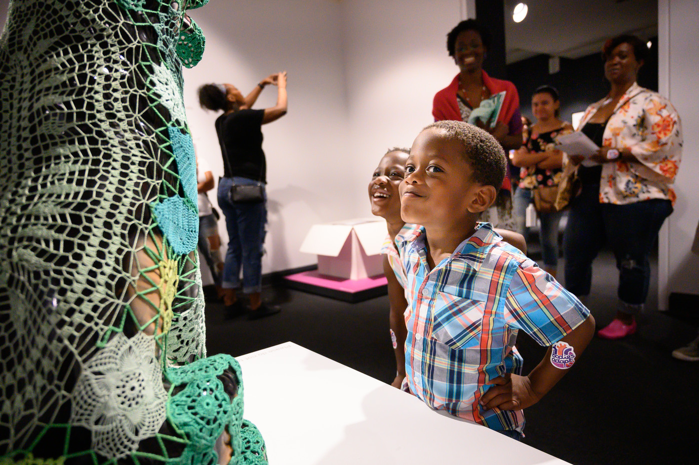 Two young children with medium skin tone stand in front of a dog sculpture covered with green crochet panels. One child stands with hands on hips, eyes turned to look directly at the viewer, and gives a slight smirk. Adult visitors in the background look at the scene with amusement.