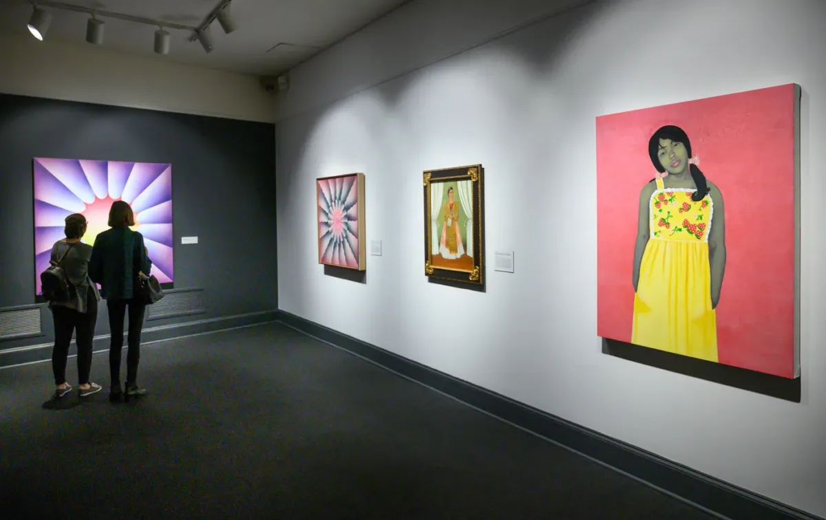 A partial view of an art gallery with four artworks on display, including a painted portrait of a young, pony-tailed medium-dark skinned girl in a yellow flowered dress against a pink background; a smaller portrait; and large abstract works with colorful shapes and gradients.