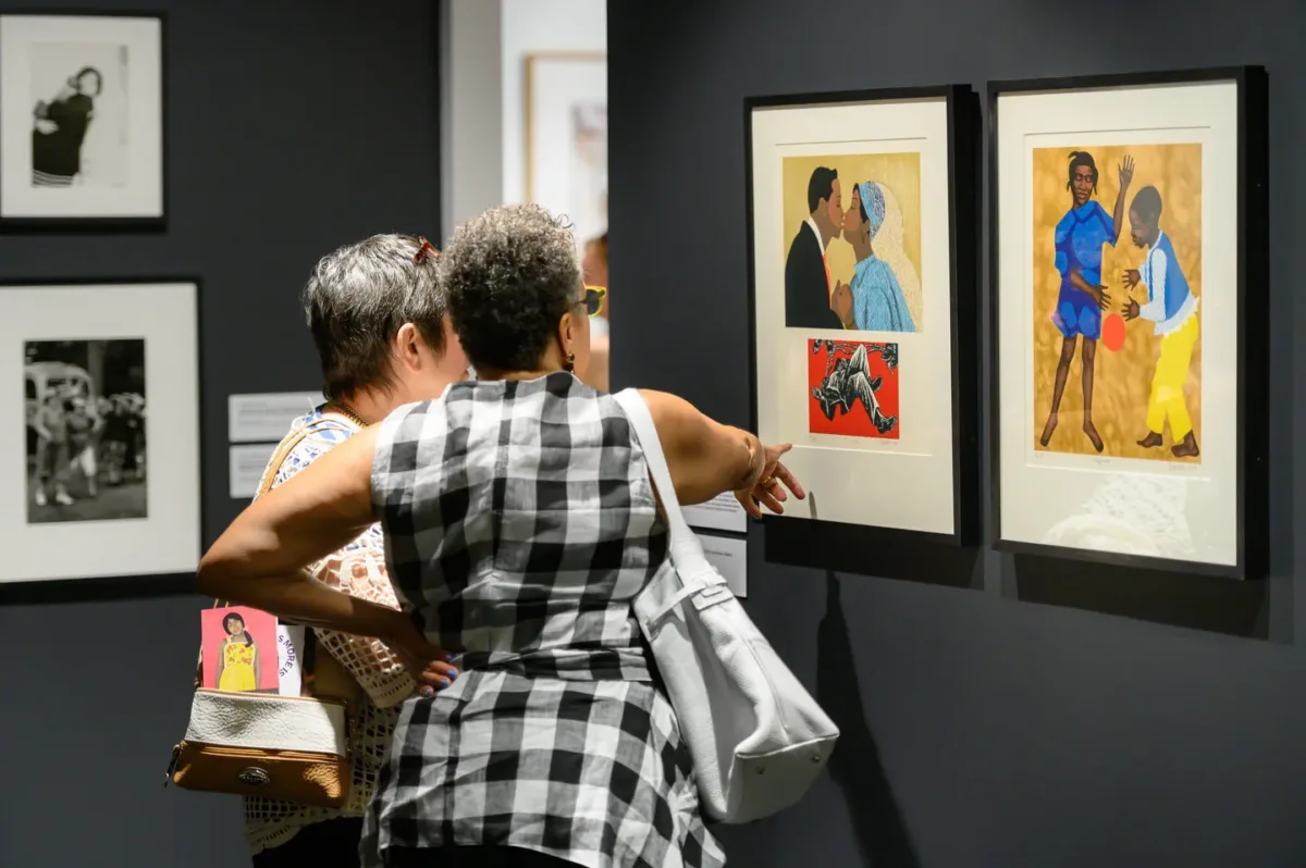 Two women with short gray hair look at brightly colored prints and point to details of the artwork.