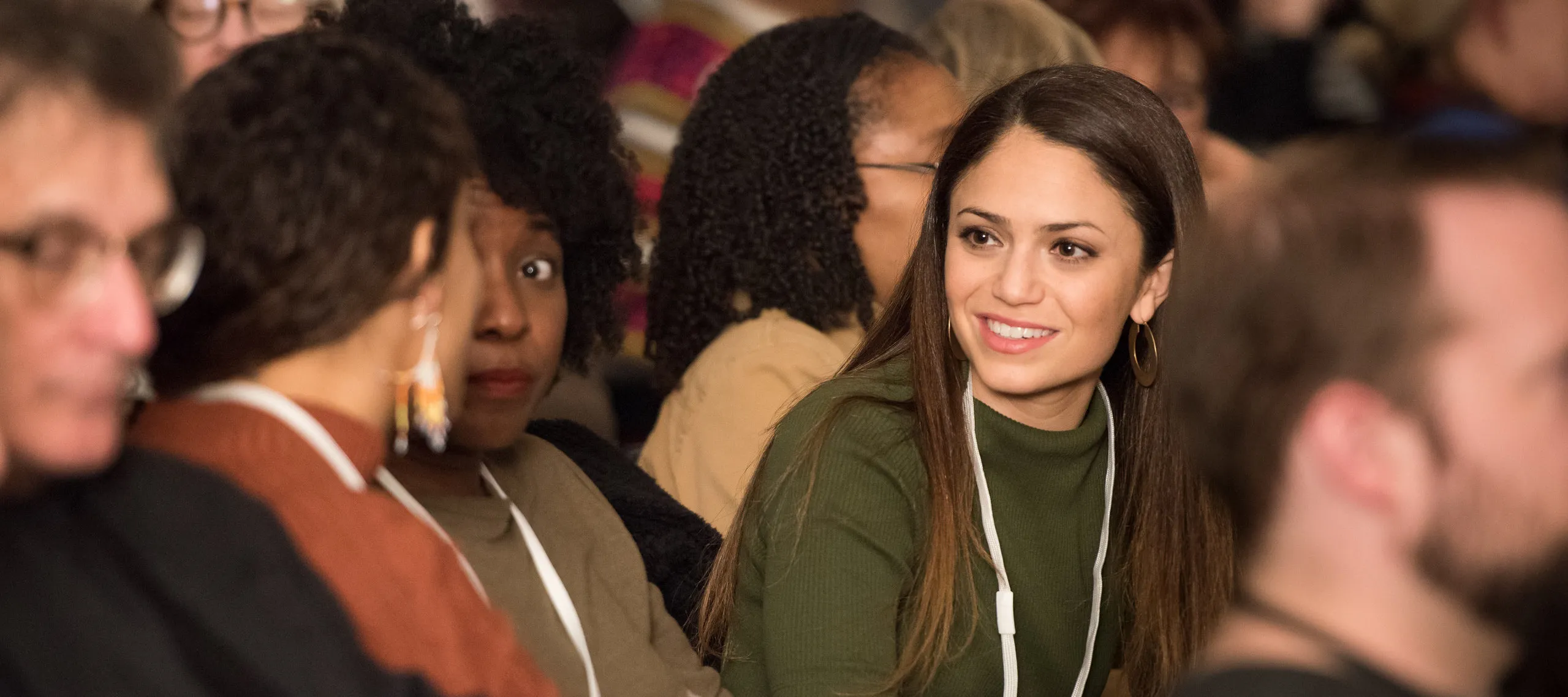 A medium light-skinned young adult smiles while listening to another adult speak a few seats over from her in a full auditorium.