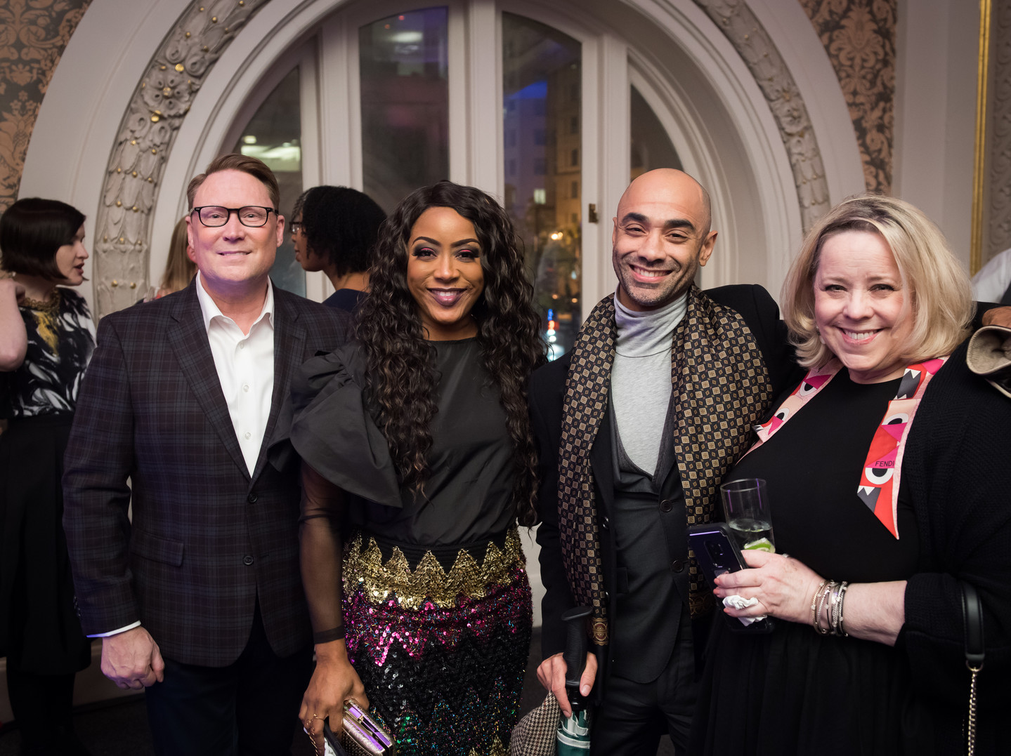 A photograph of two light-skinned people, one medium-skinned person, and one dark-skinned person at a museum event. Everyone is wearing formal attire, facing the camera, and smiling.