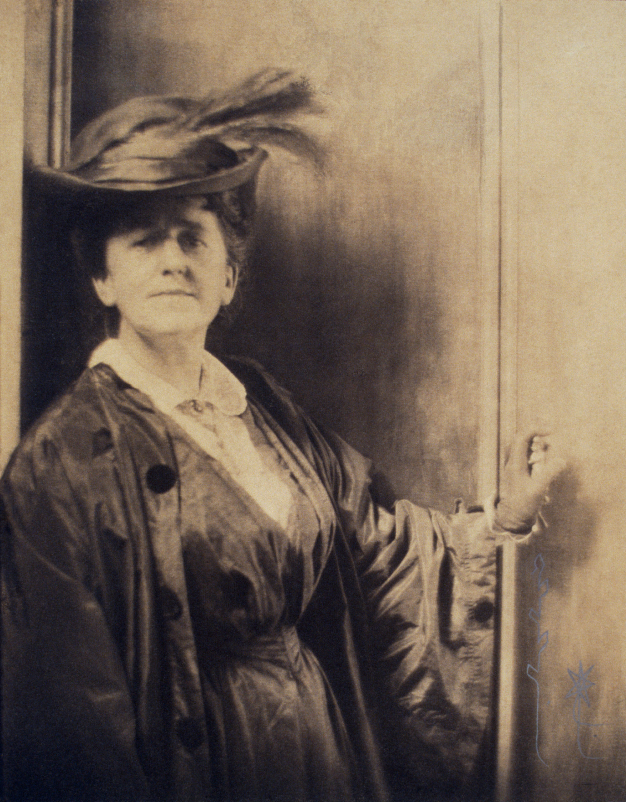 A sepia-toned photograph of a light-skinned adult woman. Standing against a wall, she dons a large black hat with a feather in the brim. She wears a dark dress with a white collared shirt and a long baggy dark coat. She places her left hand on the wall beside her.