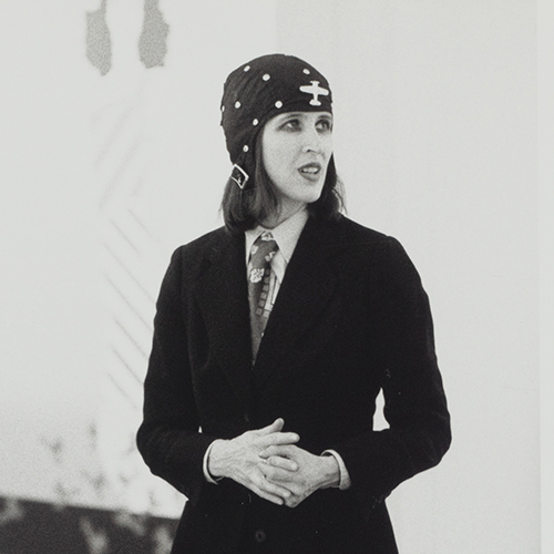 Black-and-white photograph of a light-skinned adult woman with medium-length dark hair, dark eyeliner and lipstick. She wears a dark blazer, light-color collared shirt, patterned necktie, and black cloth cap with white polkadots and airplane decal and a metal buckle.