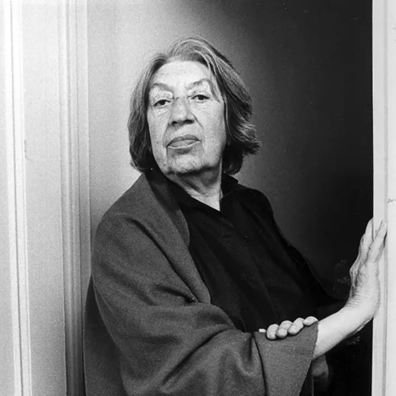 Black-and-white photograph of an older light-skinned woman with short grey and white hair. She stands in a doorway, and rests her right hand against the door frame to her left and her left hand on her right forearm. She wears a dark blouse and shawl.