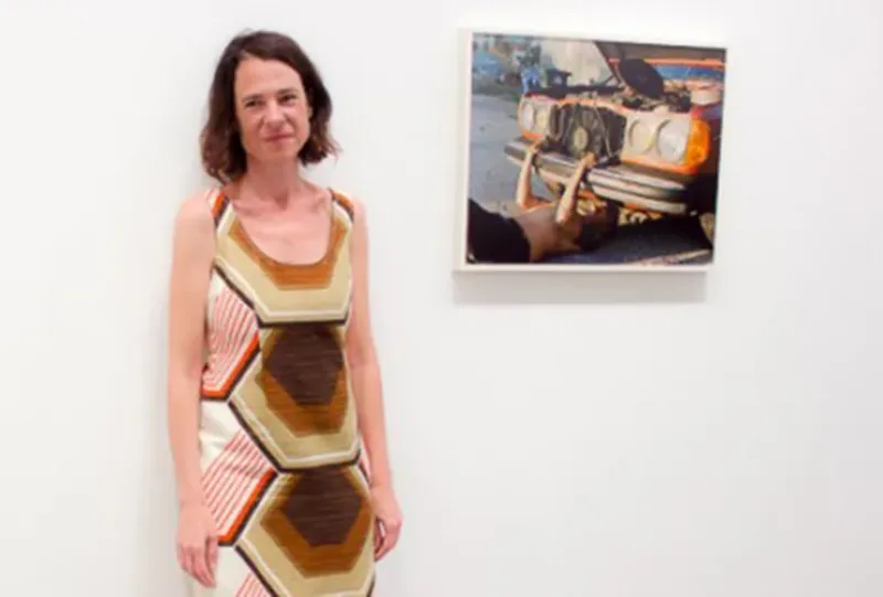 A light-skinned adult womand with bobbed brown hair stands against a white wall wearing a sleeveless dress with a colorful geometric print. On the wall behind her hangs a color photograph of a light-skinned person working outside under the hood of an old red car.