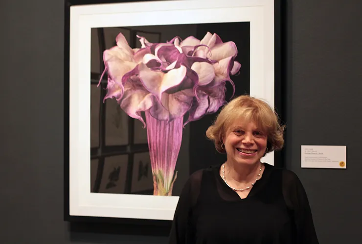 A light-skinned, adult woman with short, blonde hair grins in front of one of her photographs depicting a blossoming, purple datura flower against a black background.