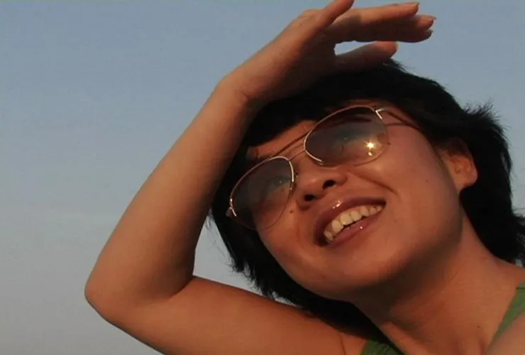 A color photograph of a medium-brown skinned woman, wearing aviator sunglasses against a blue background. She smiles as she gazes upwards, shading her eyes with her hand. The sun glints on the sunglasses and her glossy lips.